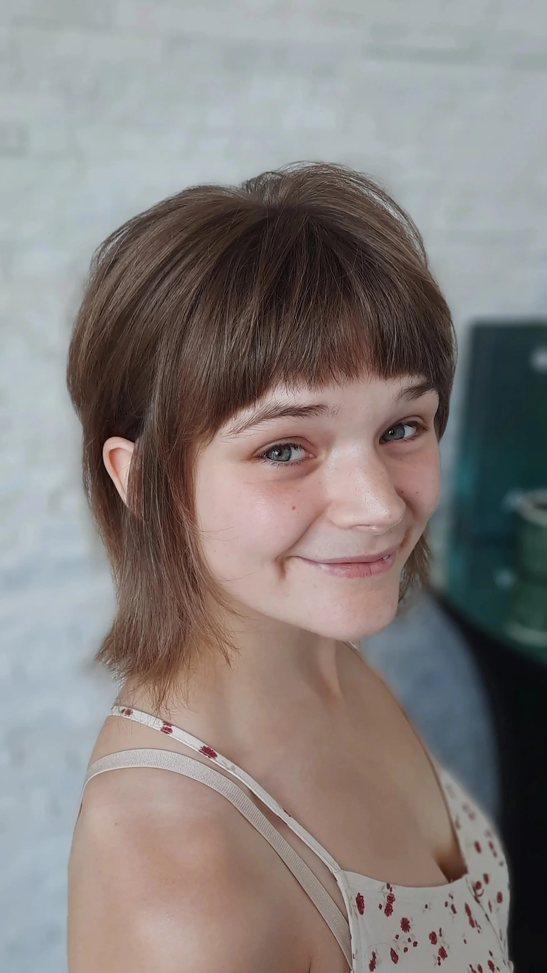 A young woman with short brown hair, light skin, and blue eyes, smiling while looking at the camera. She is wearing a sleeveless dress with a floral pattern.