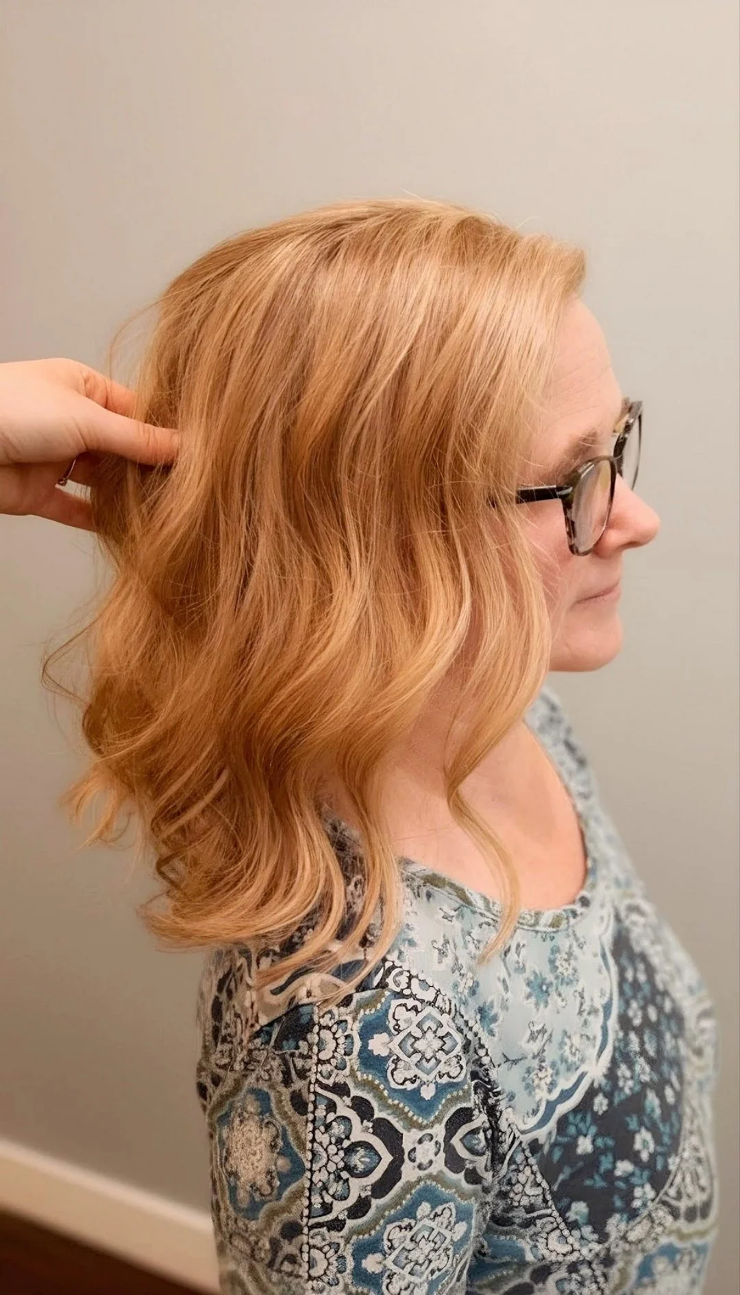 A woman with shoulder-length wavy, light red hair wearing glasses and a patterned blouse, standing against a plain wall.