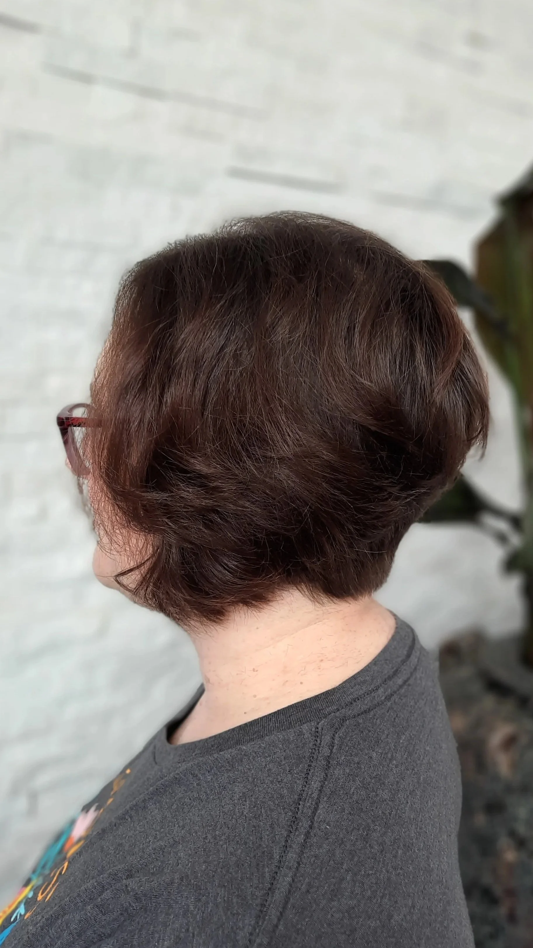 Back view of a woman with short, reddish-brown hair, wearing glasses and a dark gray t-shirt, standing indoors against a light-colored wall with some greenery nearby.
