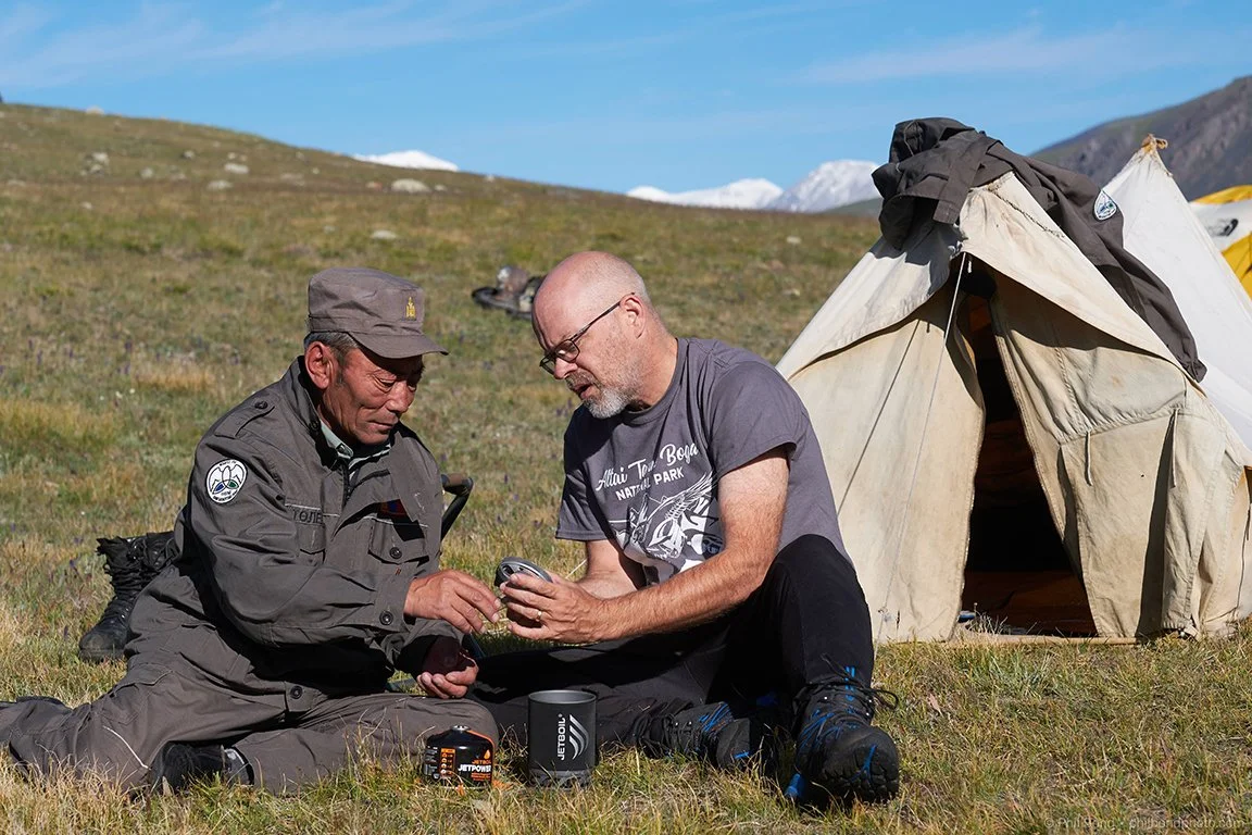 Tom Medema sitting in a mountain meadow with a ranger talking about how a jetboil system works.