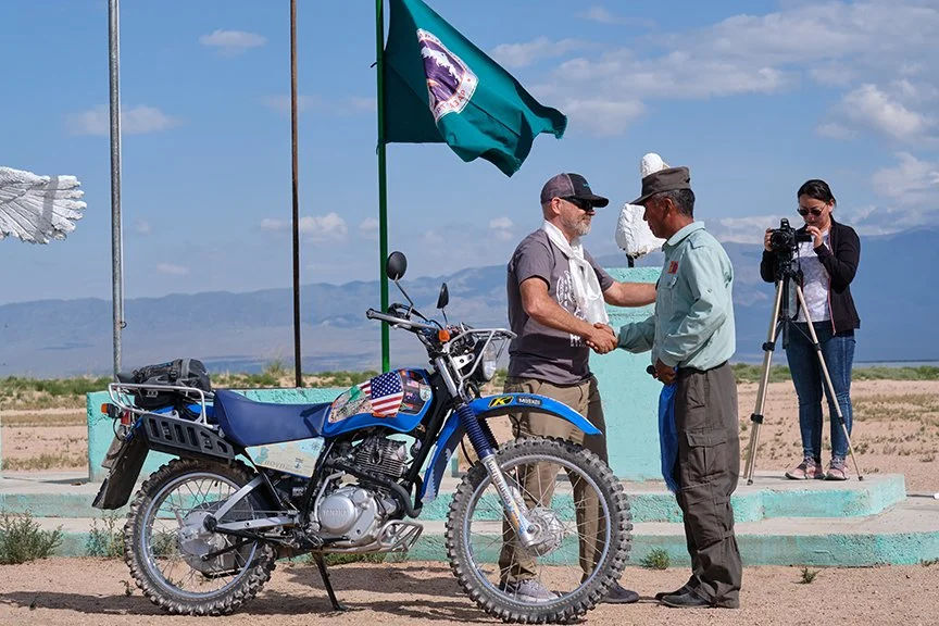Tom Medema shakes hands with a Mongolian Park Ranger with a motorcycle he is donating to that park ranger at an outdoor public ceremony