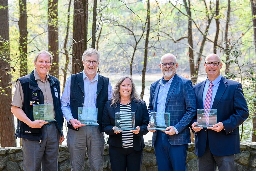A group of five award recipients pose with their glass plaques in a wooded amphitheater.