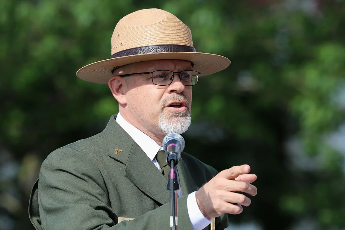 Tom Medema in a formal park ranger uniform points and speaks into a microphone at a public event in Anniston Alabama