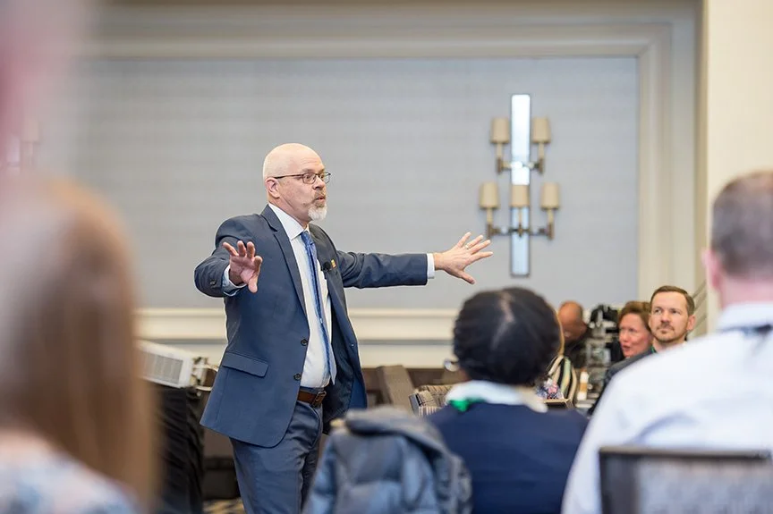 Tom Medema in a blue suit giving a presentation or speech to an audience in a conference room.