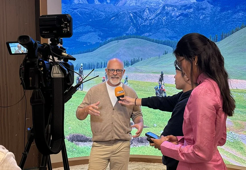 Tom Medema being interviewed by Mongolian television in front of a mountain landscape with motorcycles riding through