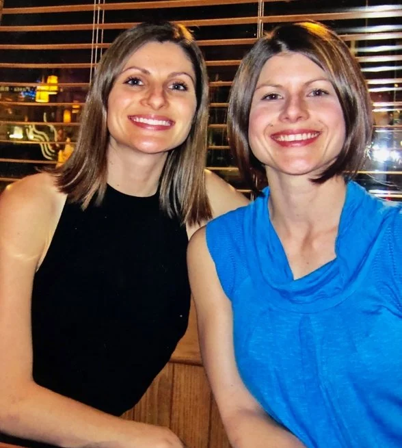 Two sisters smiling, sitting together indoors in front of a window with wooden blinds, smiling at the camera.