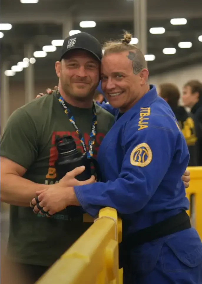 Two men smiling and hugging; one in a green t-shirt and black cap, the other in a blue martial arts uniform with yellow insignia and the name "TULIA" on the sleeve; they are at a sporting event with yellow barricades and other people in the background.