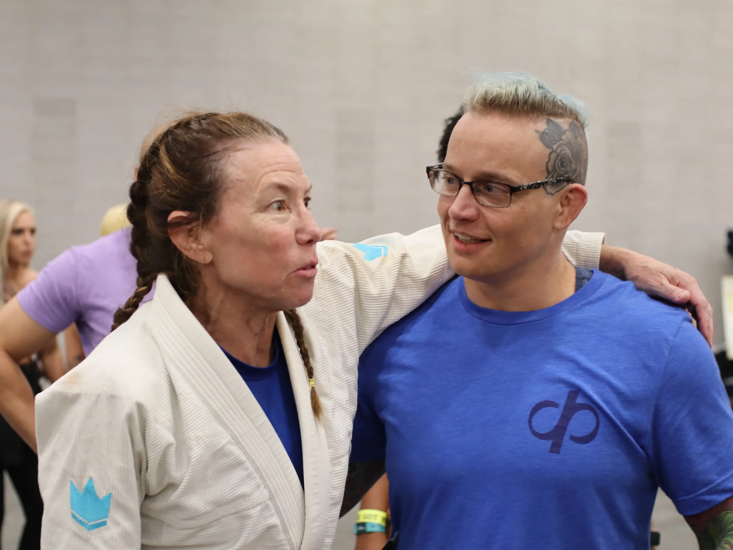 A woman and a man are sharing a friendly moment, smiling and looking into each other's eyes, with their arms around each other's shoulders.