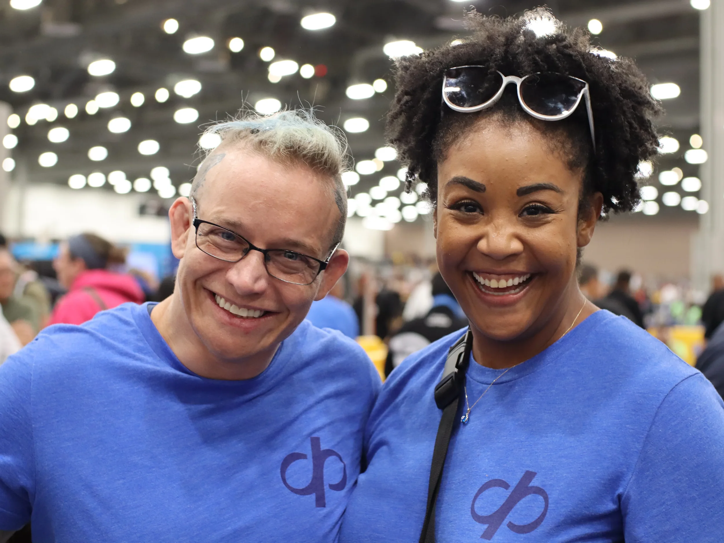 Two smiling people, a man with glasses and dyed hair, and a woman wearing sunglasses on her head, at an indoor event or convention.