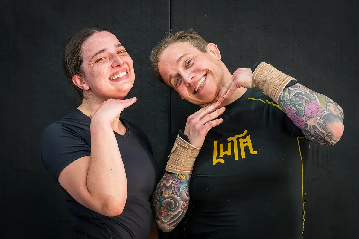 Two women smiling and posing against a black background, one with short dark hair and the other with short light brown hair, both wearing athletic clothing.