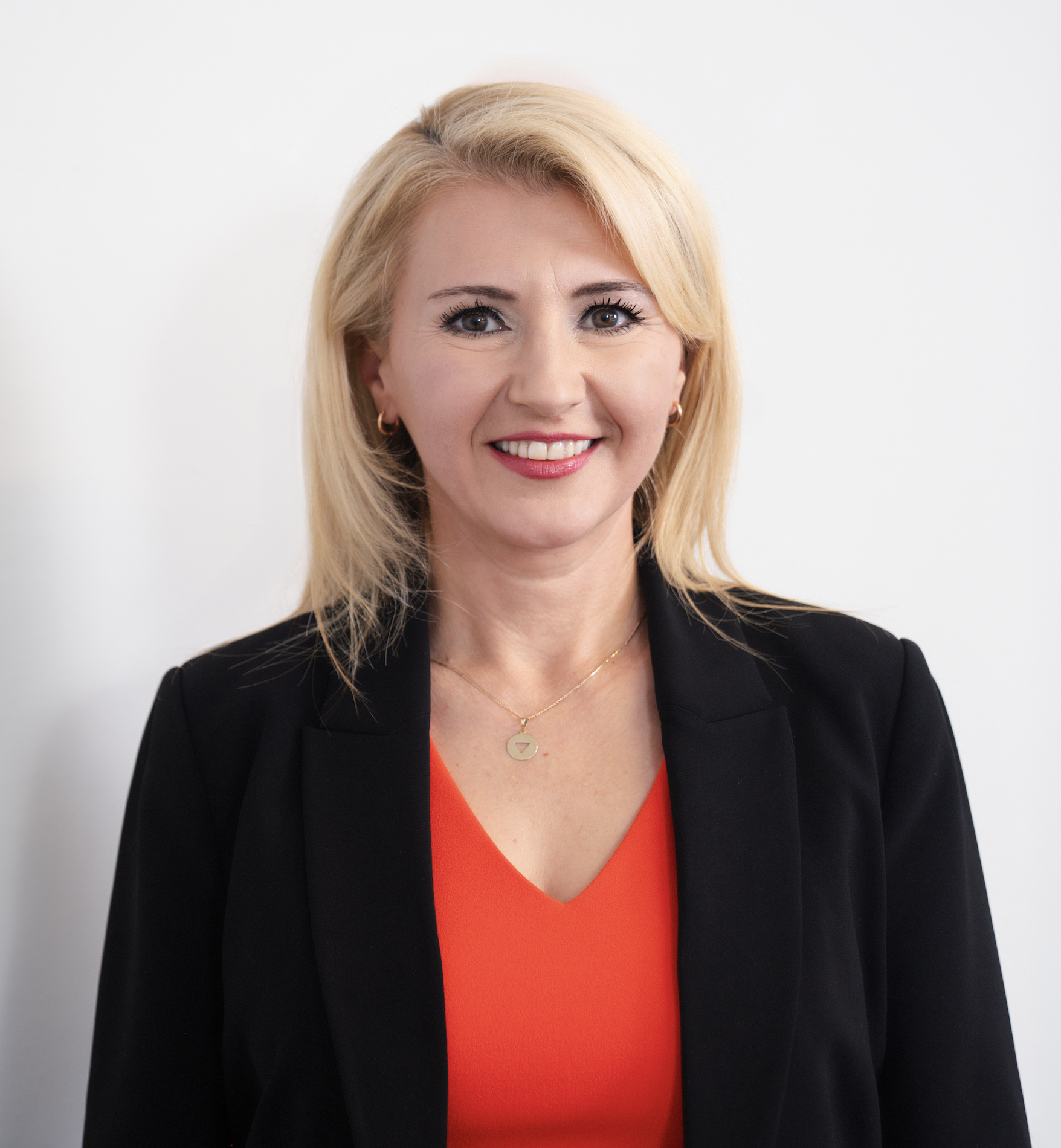 A professional woman with blonde hair, wearing a black blazer over a red top, accessorized with a gold necklace and earrings, smiling against a plain white background.