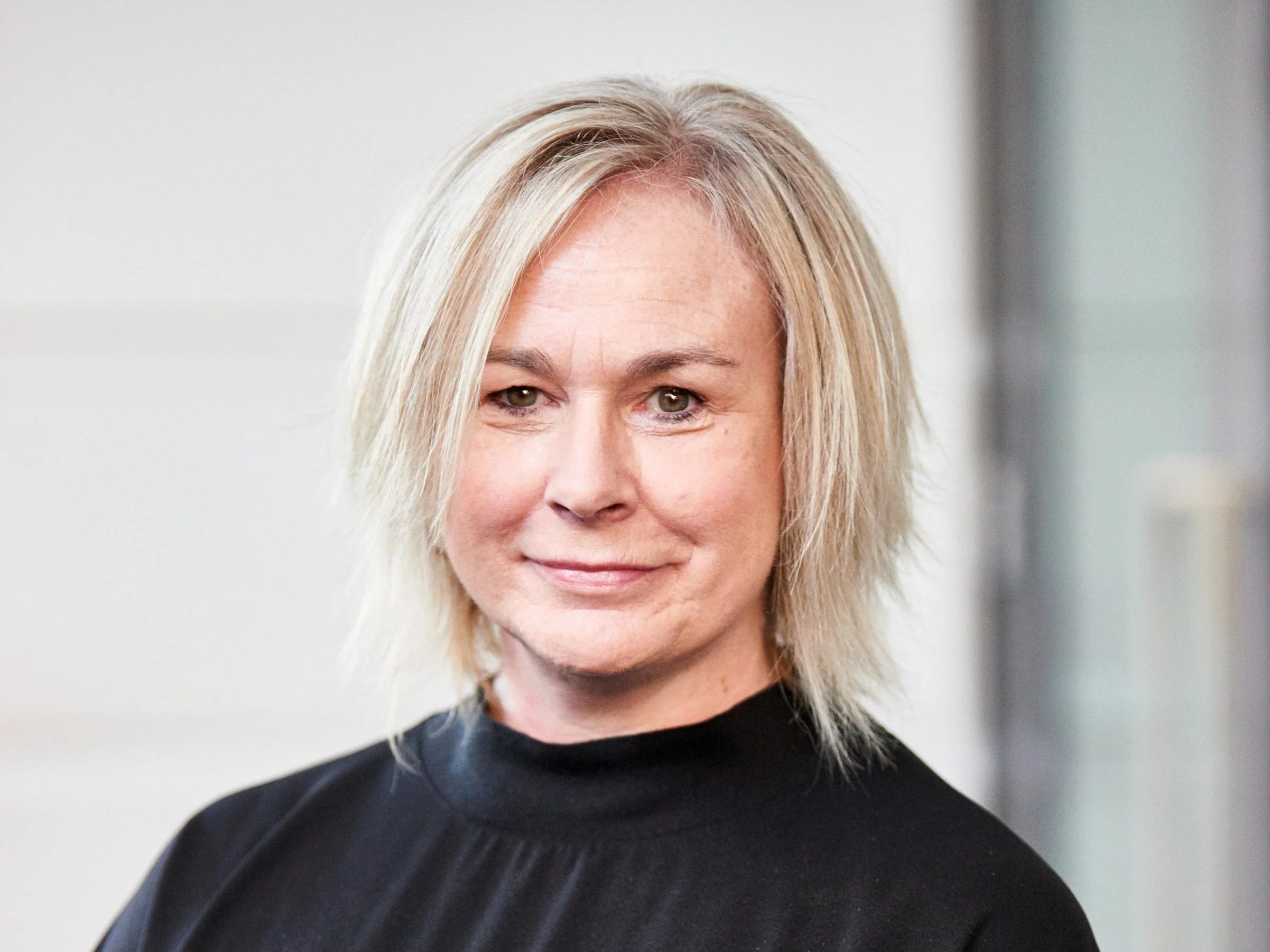 A middle-aged woman with blonde hair and green eyes, wearing a black top, smiling softly in front of a plain light-colored background.