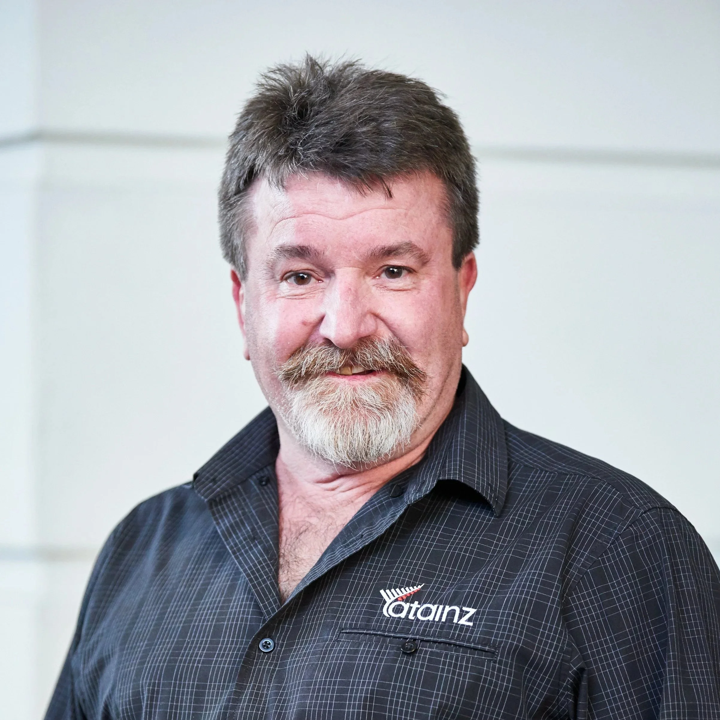 Close-up of a man with a full beard and mustache, wearing a black plaid shirt with a logo that reads 'CATainz'.