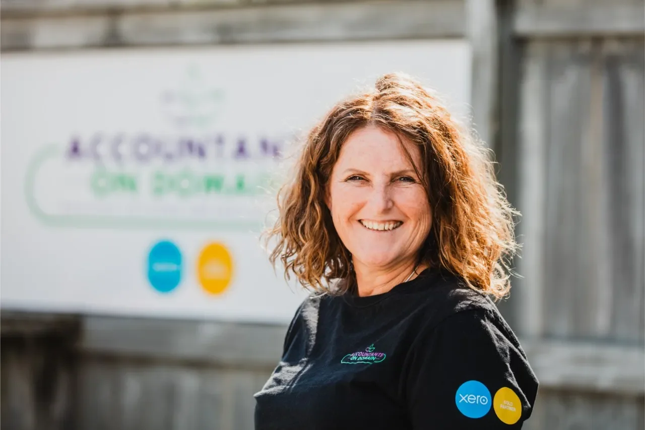 A smiling woman with curly red hair in front of a blurred outdoor background with a sign that reads "Accountants Can Own." She is wearing a black shirt with a blue sticker that says "Xero" and a yellow sticker that says "Gold Partner."