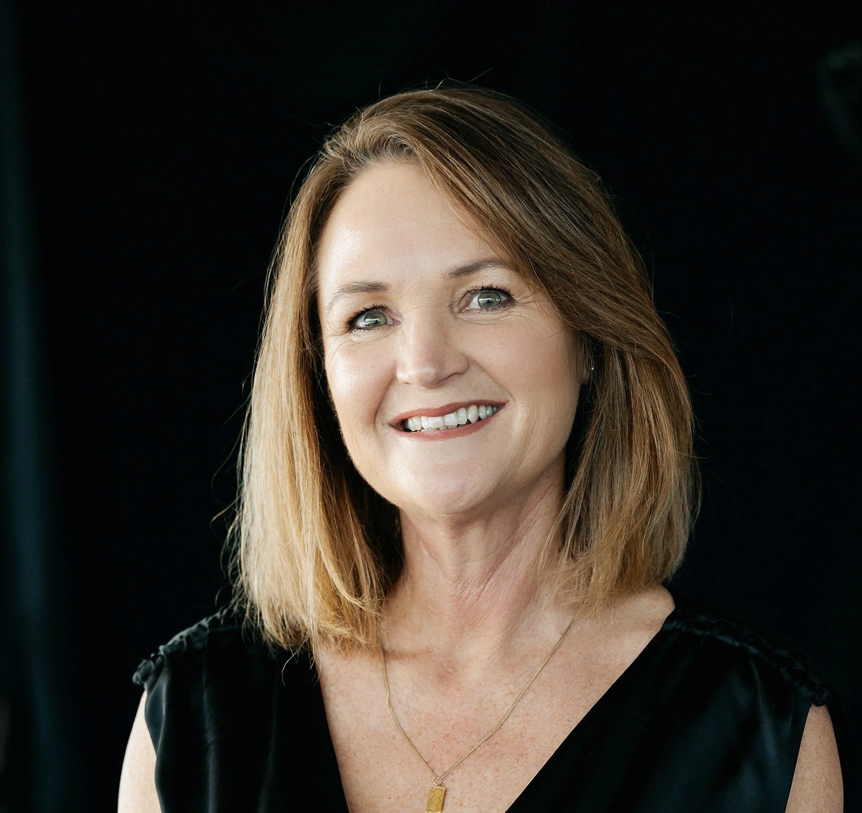 A woman with long ginger hair, light skin, blue eyes, and a bright smile, wearing a black top and a gold necklace, against a black background.