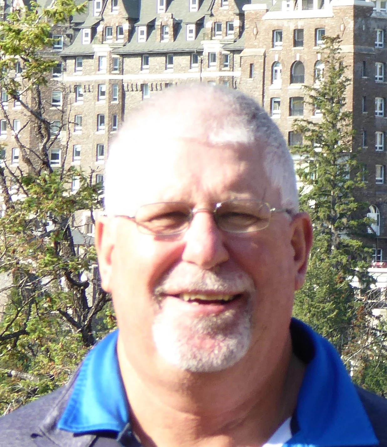 A smiling older man with glasses and a beard outdoors, with buildings and trees in the background.