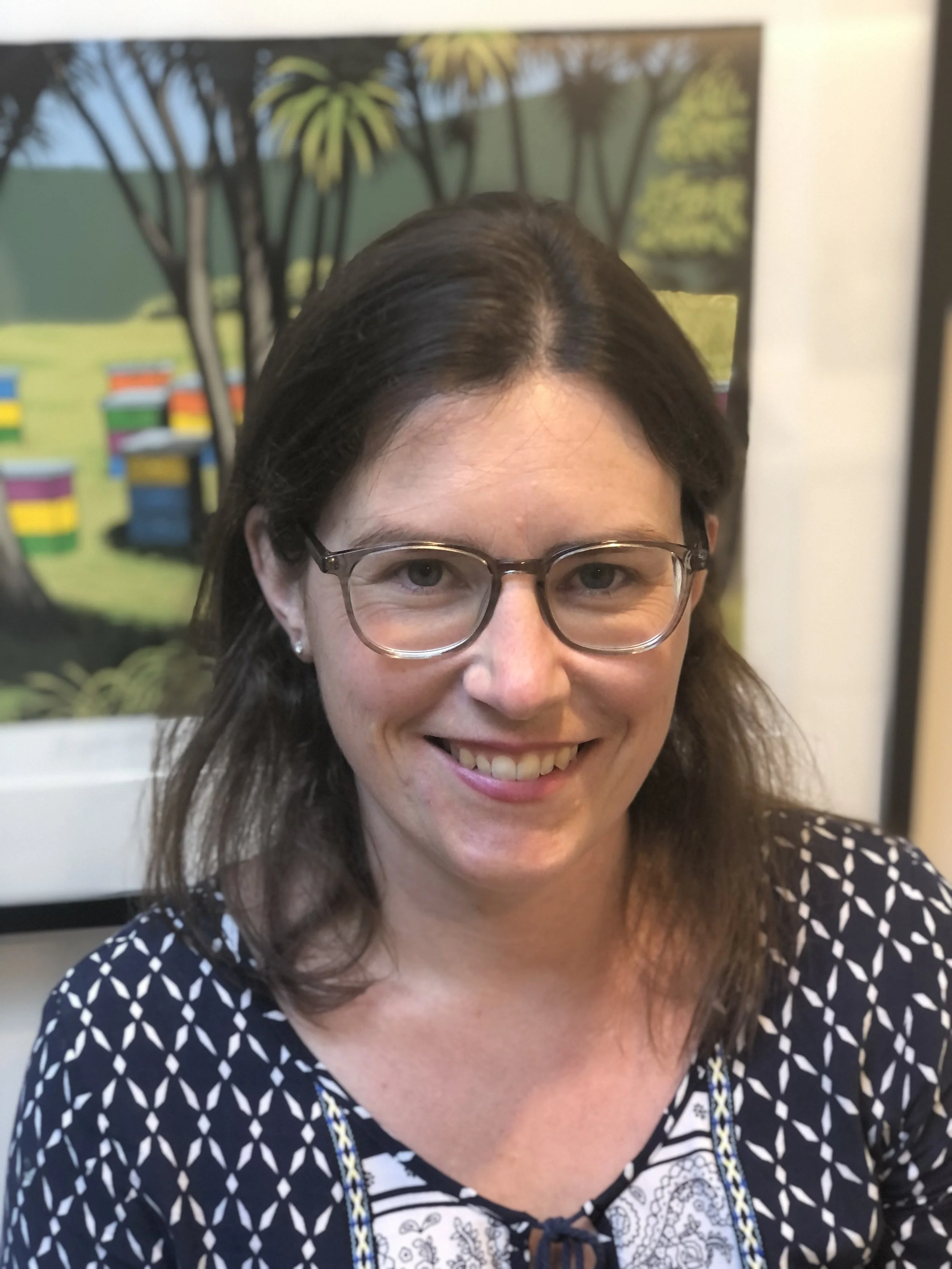 A woman with brown hair and glasses smiling, wearing a black dress with white geometric pattern, in front of a colorful outdoor scene painting.