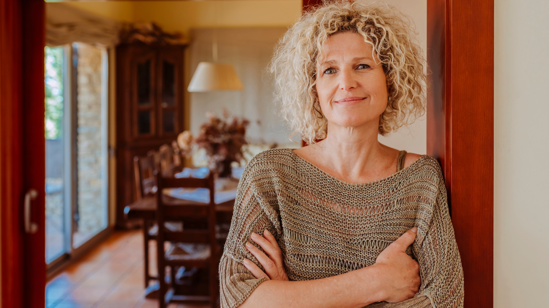 An older woman with curly blonde hair stands in a doorway inside a house, smiling softly and crossing her arms, wearing a loose, knit, beige sweater.