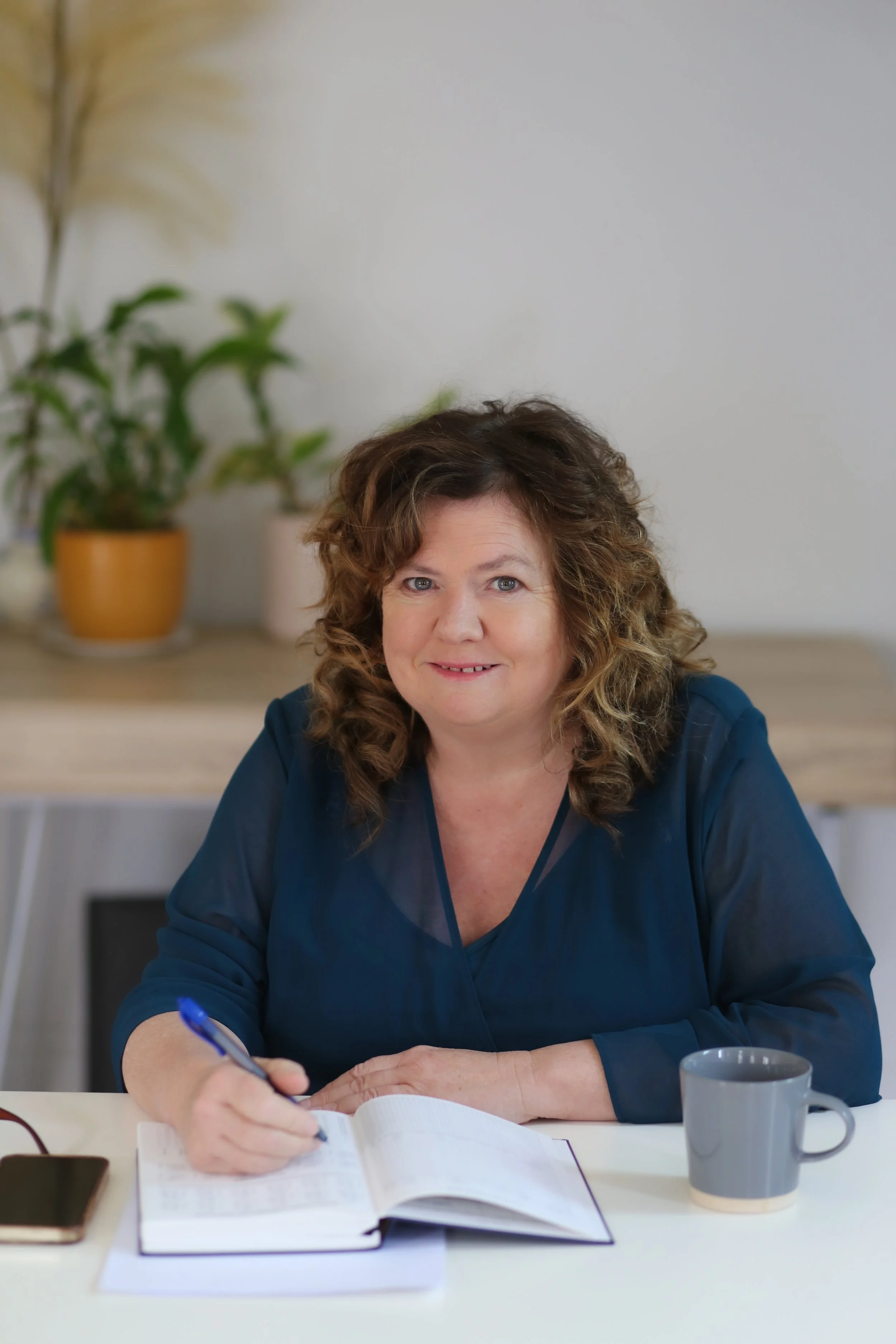 A woman with curly brown hair in a blue blouse sitting at a table, writing in a notebook, with a cup, a potted plant, and a smartphone on the table.