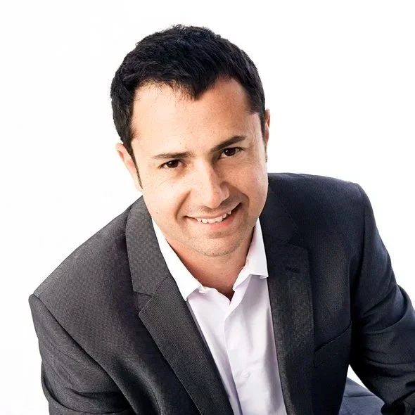 A man smiling, wearing a dark gray suit and white shirt, with dark hair, against a white background.