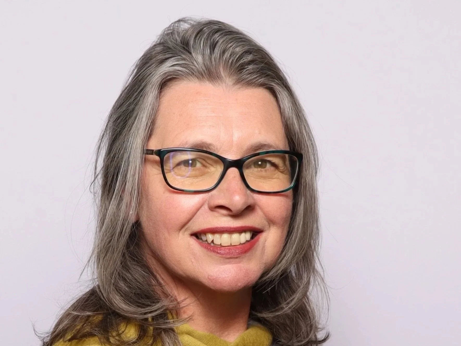 A middle-aged woman with gray hair, glasses, and a yellow sweater smiling in front of a plain white background.
