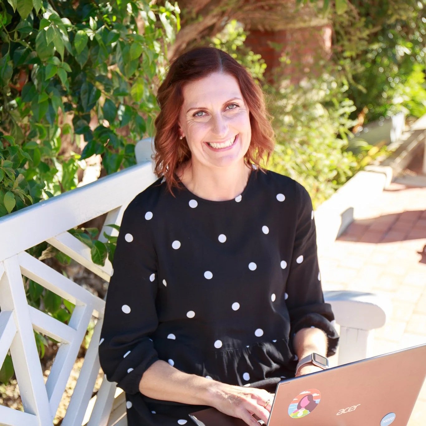 A woman with red hair smiling while sitting on a white park bench with greenery in the background, working on a laptop.