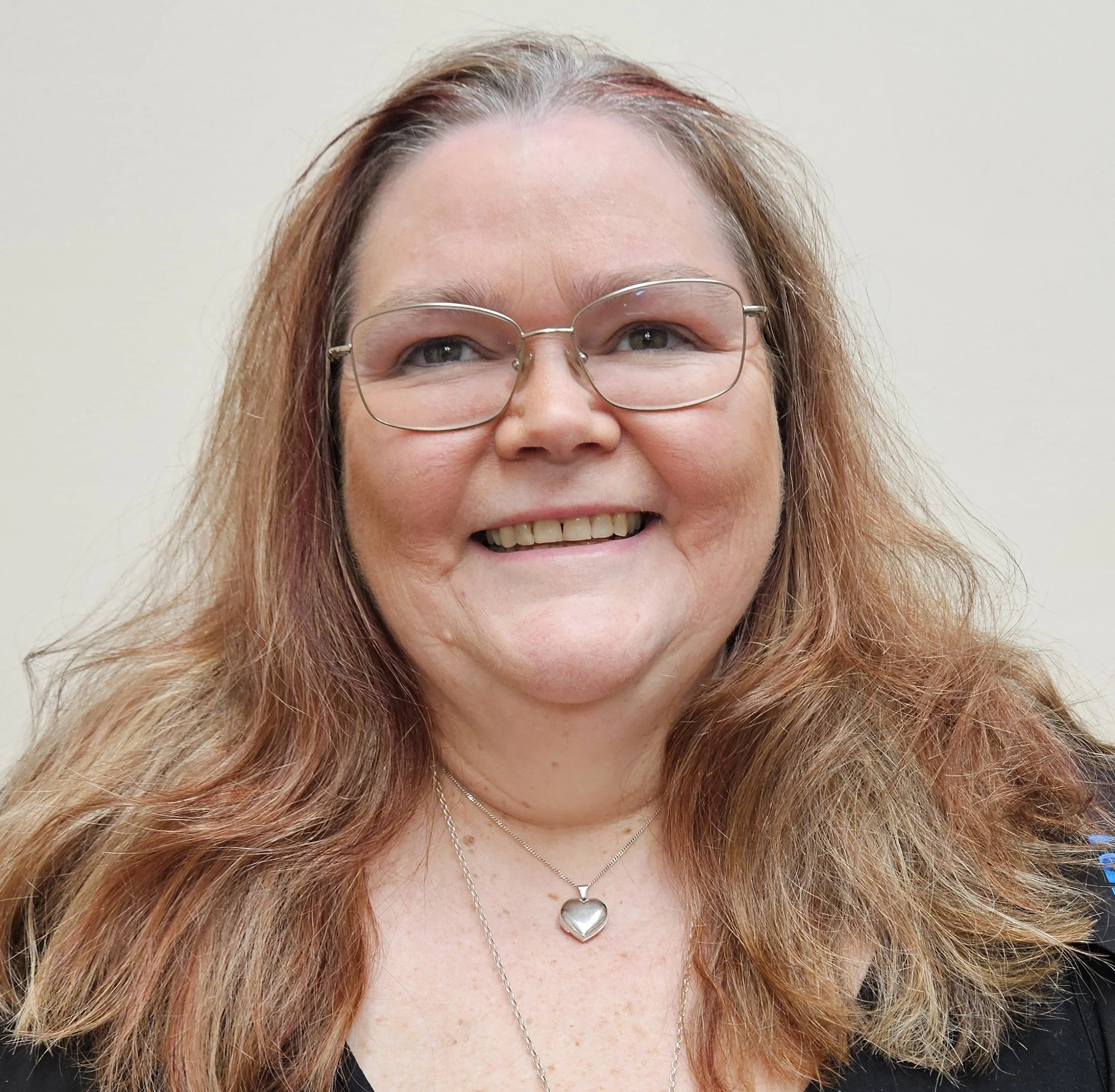 A woman with red hair, glasses, and a heart-shaped necklace smiling in front of a plain off-white wall.