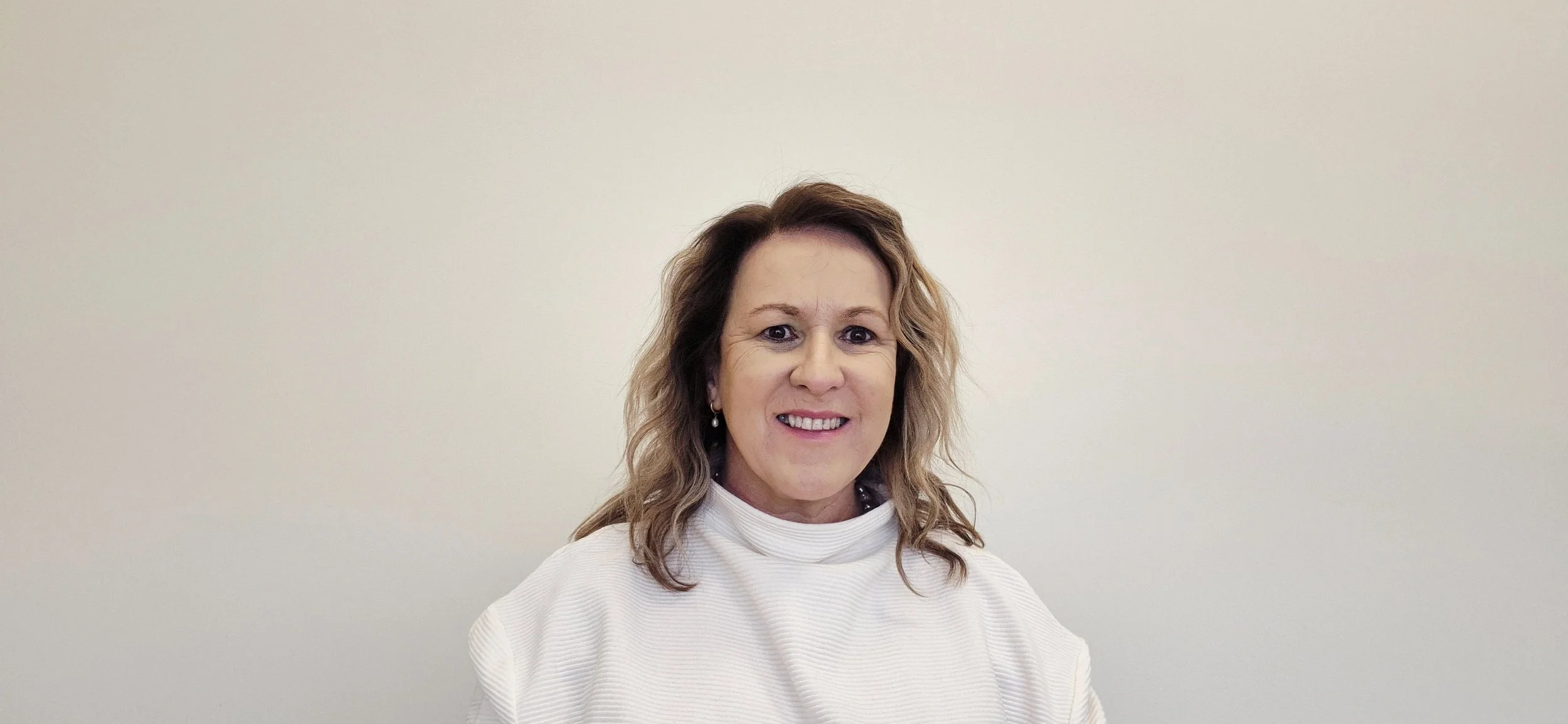 A woman with wavy, shoulder-length hair smiling at the camera, wearing a white top, against a plain light-colored wall.