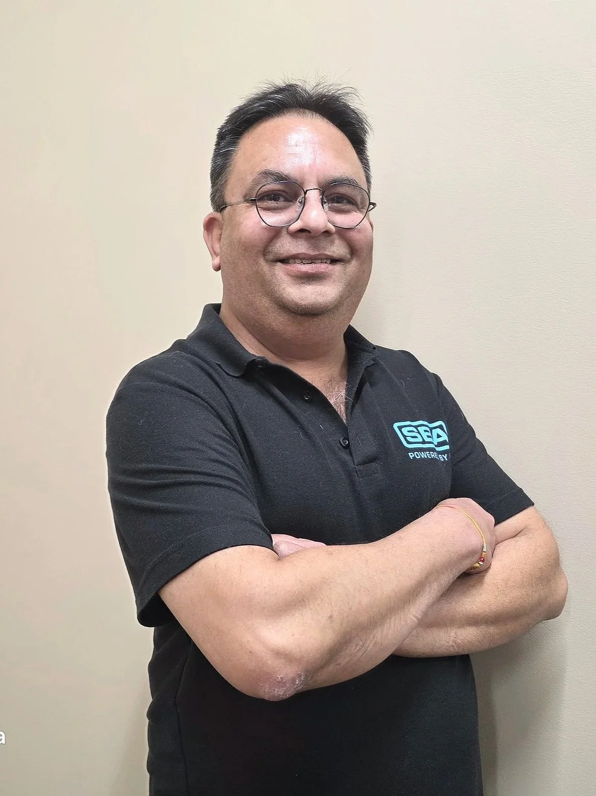 A man with glasses and dark hair, smiling with arms crossed, standing in front of a plain beige wall, wearing a black polo shirt with a logo on the chest.