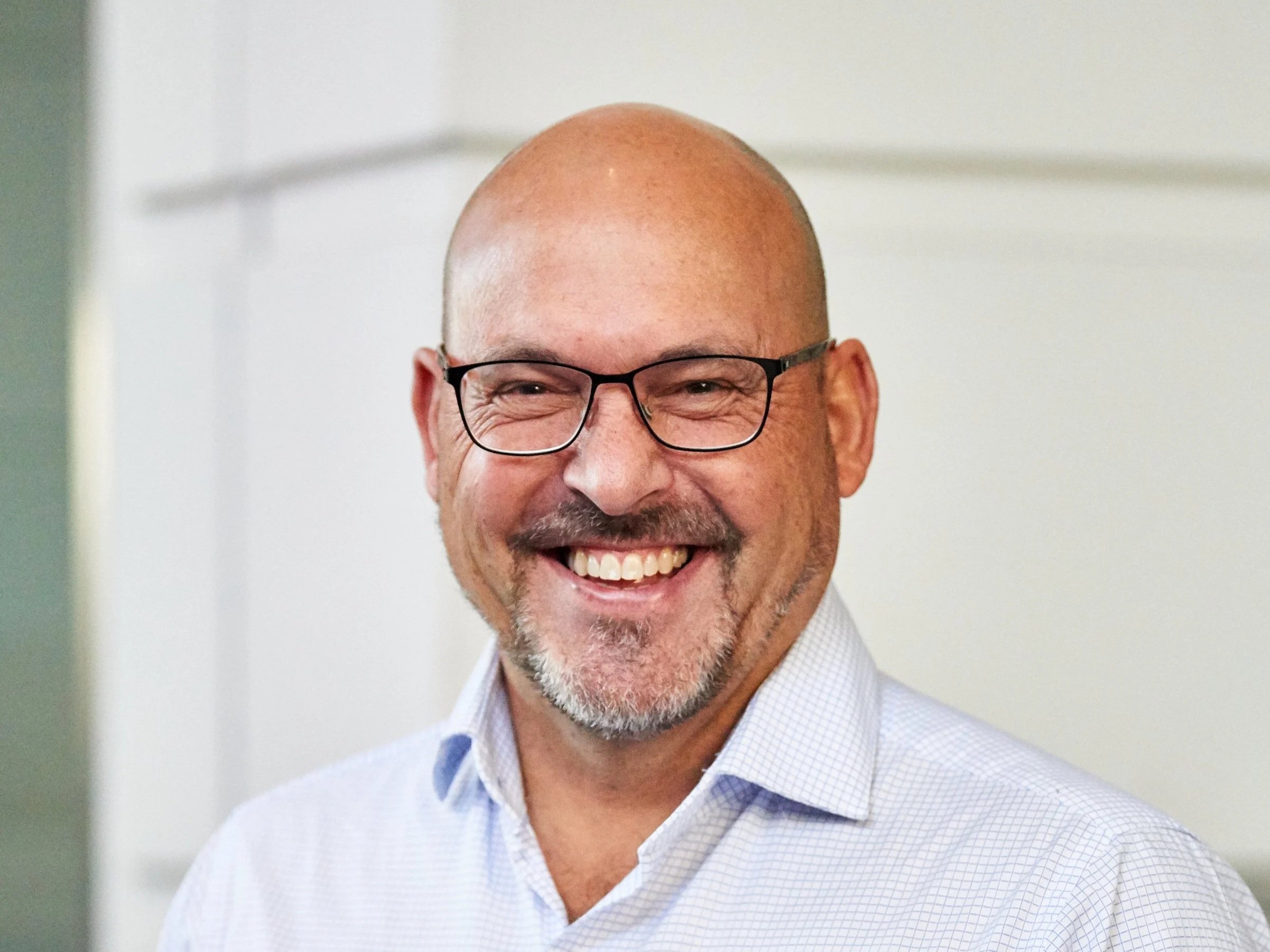 A smiling bald man with glasses and a gray beard, wearing a light blue checkered shirt.