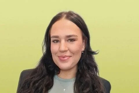 A young woman with dark hair and a gray top, smiling in front of a light green background.