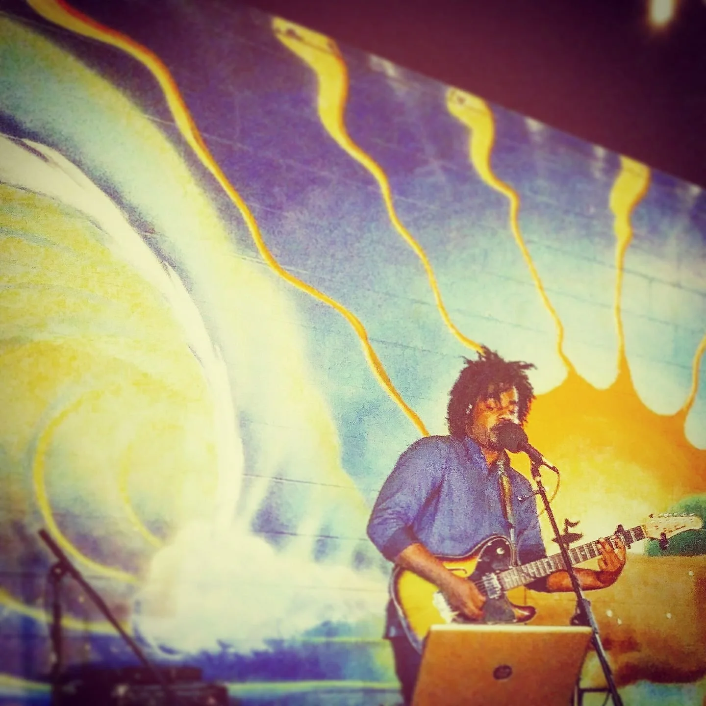 A musician with curly hair and a beard performing on stage, playing an electric guitar and singing into a microphone. The background features colorful abstract artwork with yellow and purple wave patterns.