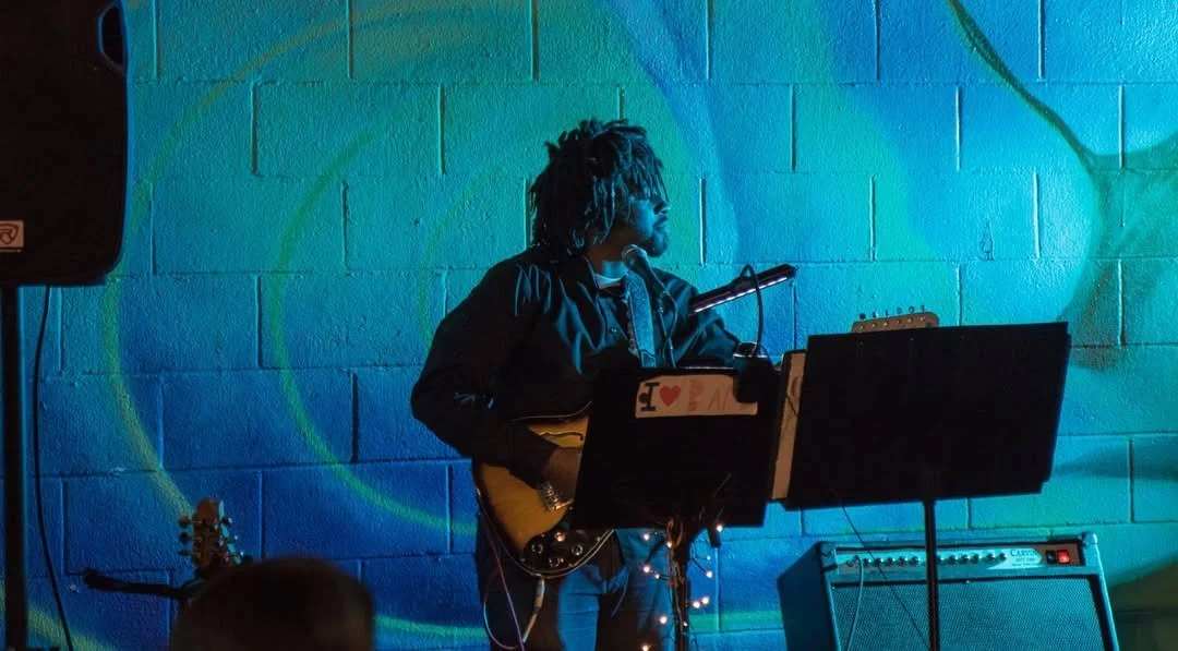 Musician with dreadlocks playing guitar on stage with blue and green lighting, in front of a music stand and amplifier, against a brick wall.