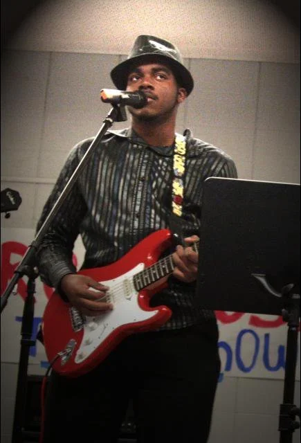 A man wearing a black hat and dark striped shirt is playing an electric guitar and singing into a microphone in a studio or performance space.