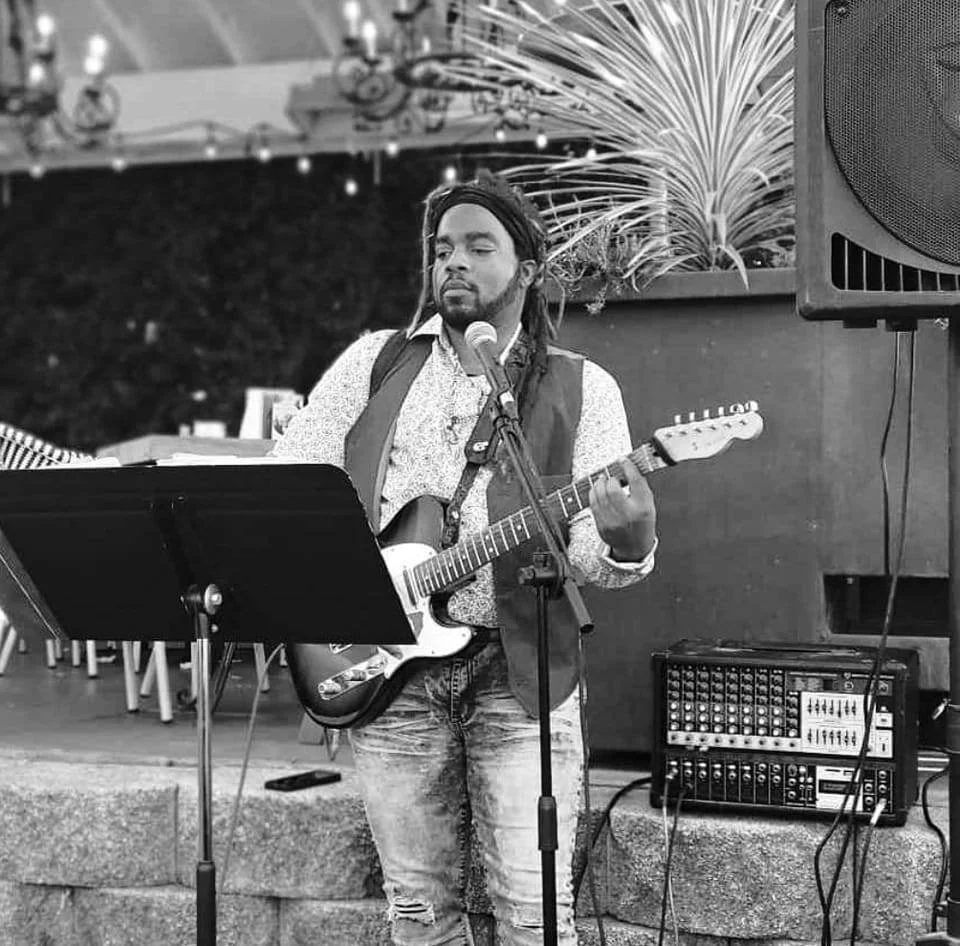 A man with dreadlocks, wearing a vest and jeans, playing an electric guitar and singing into a microphone at an indoor or outdoor event.