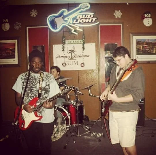 Three young musicians performing on stage with guitars and drums, decorated with holiday snowflakes and Christmas-themed signs, under a Bud Light logo sign.