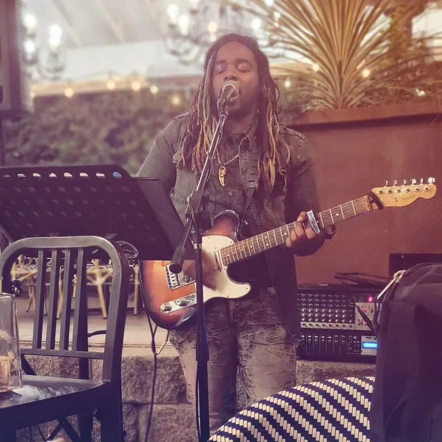 A man with long dreadlocks singing into a microphone and playing an electric guitar during a live performance at an outdoor venue.