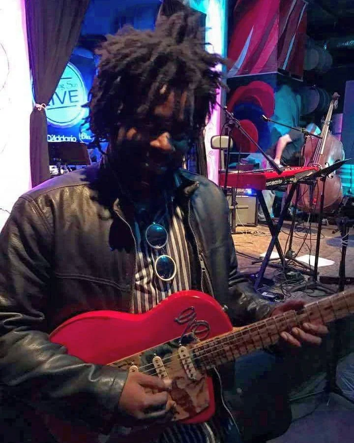 A musician with dreadlocks playing a red electric guitar on stage with other instruments in the background.