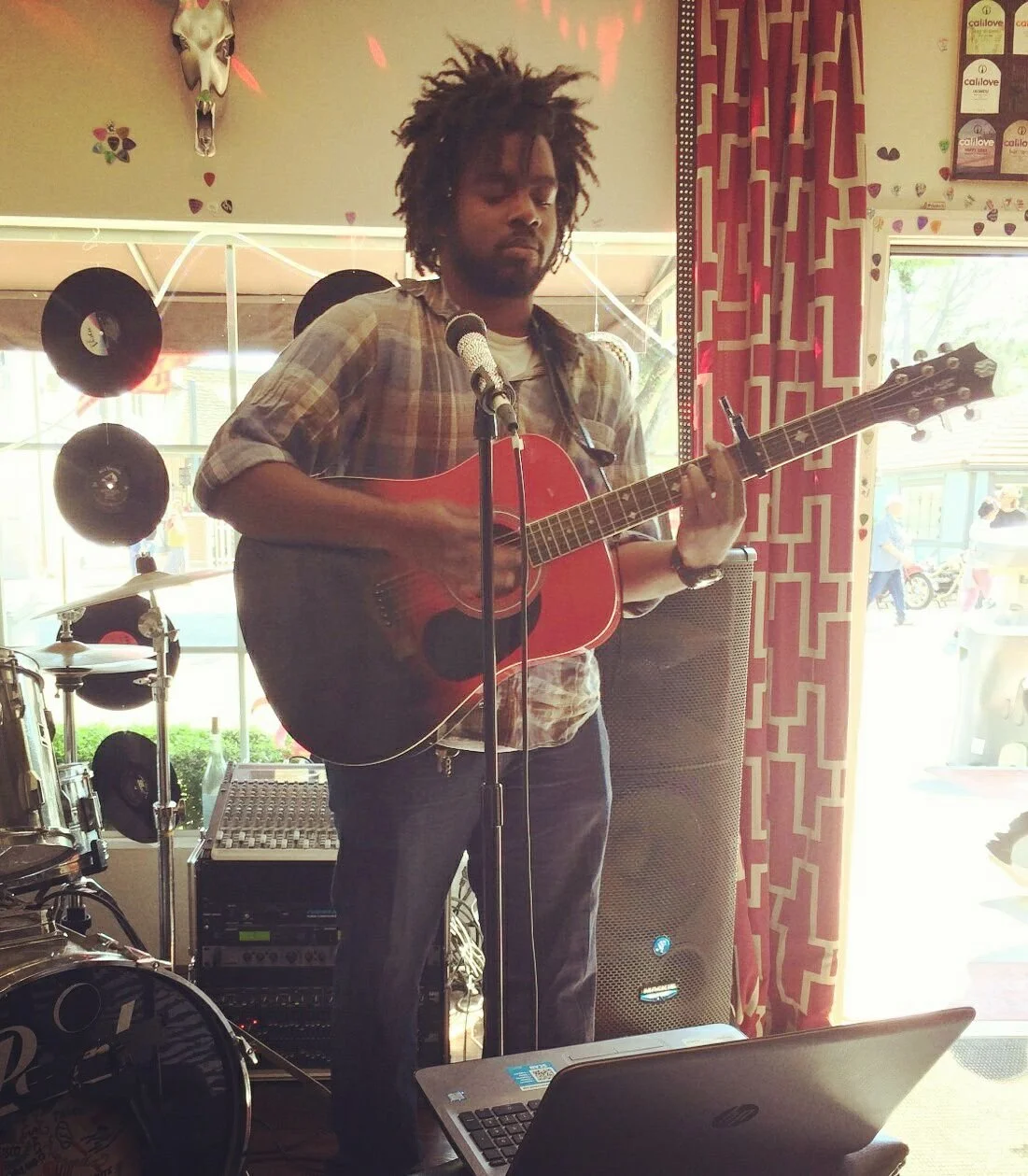 A man with dreadlocks is playing an acoustic guitar and singing into a microphone inside a room decorated with vinyl records and a deer skull.