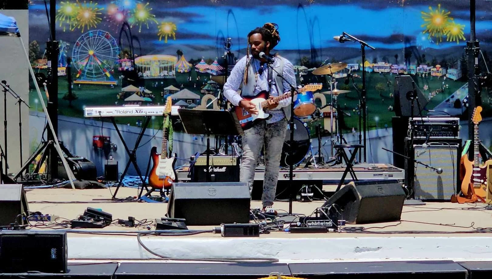 Musician with dreadlocks playing an electric guitar on stage with a colorful fairground backdrop including a Ferris wheel and tents.