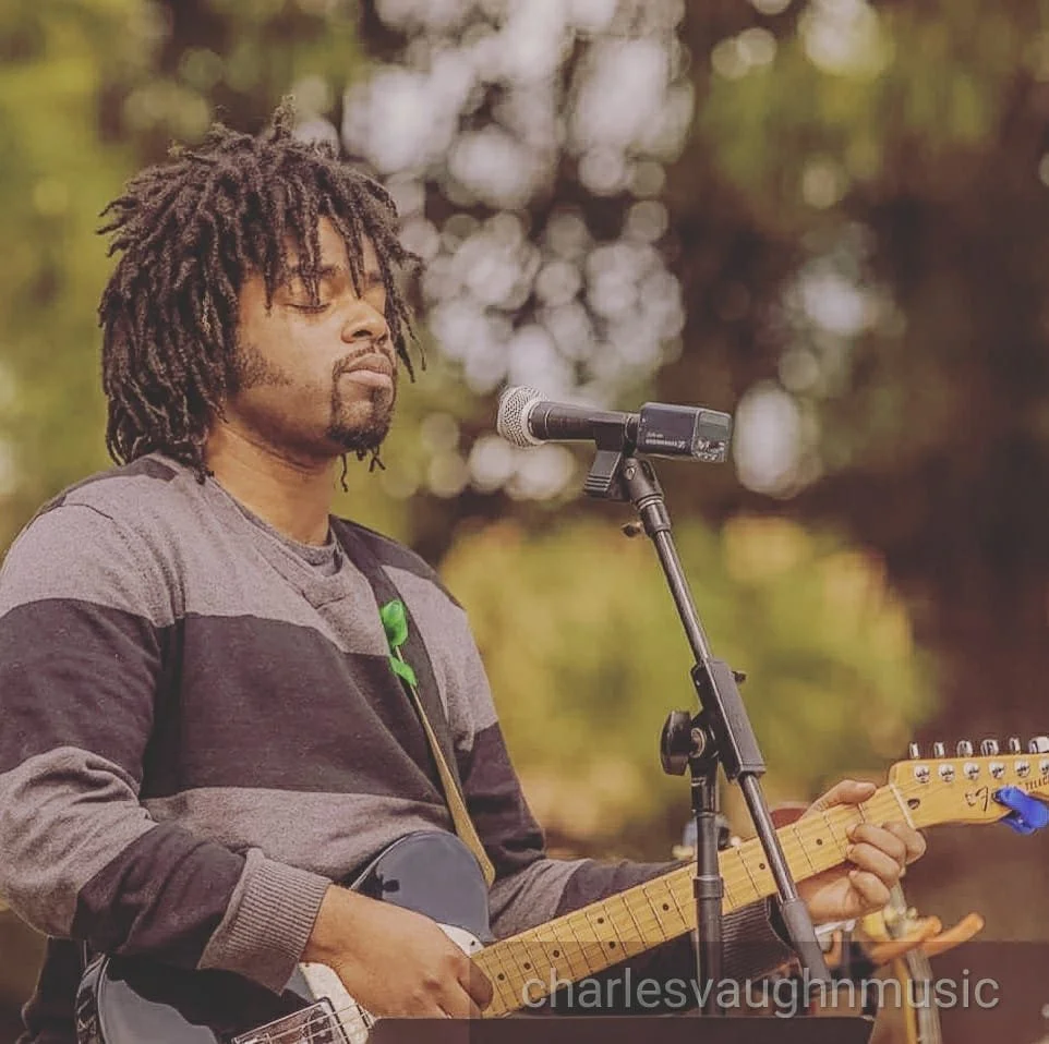 A man with dreadlocks playing an electric guitar and singing into a microphone outdoors with a blurred background.