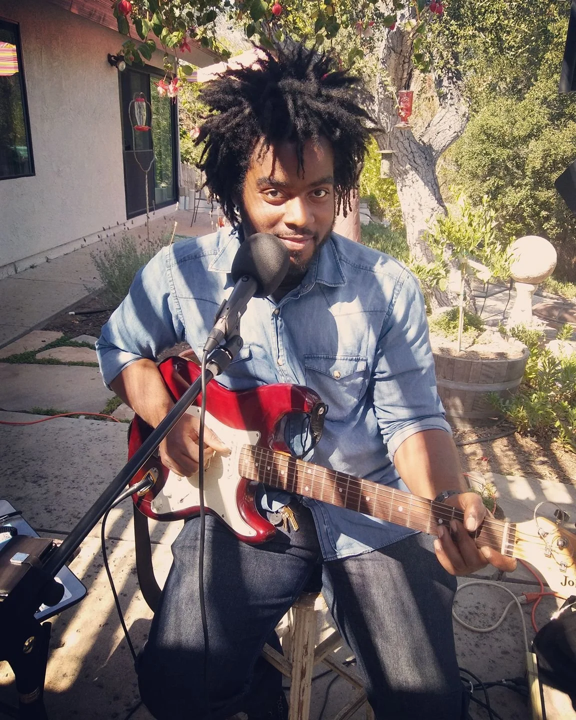 A man sitting outdoors playing an electric guitar in front of a microphone, with a garden and a house in the background.