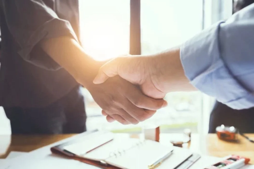 A close-up of a handshake between two individuals in a professional setting, with a window in the background allowing natural light.