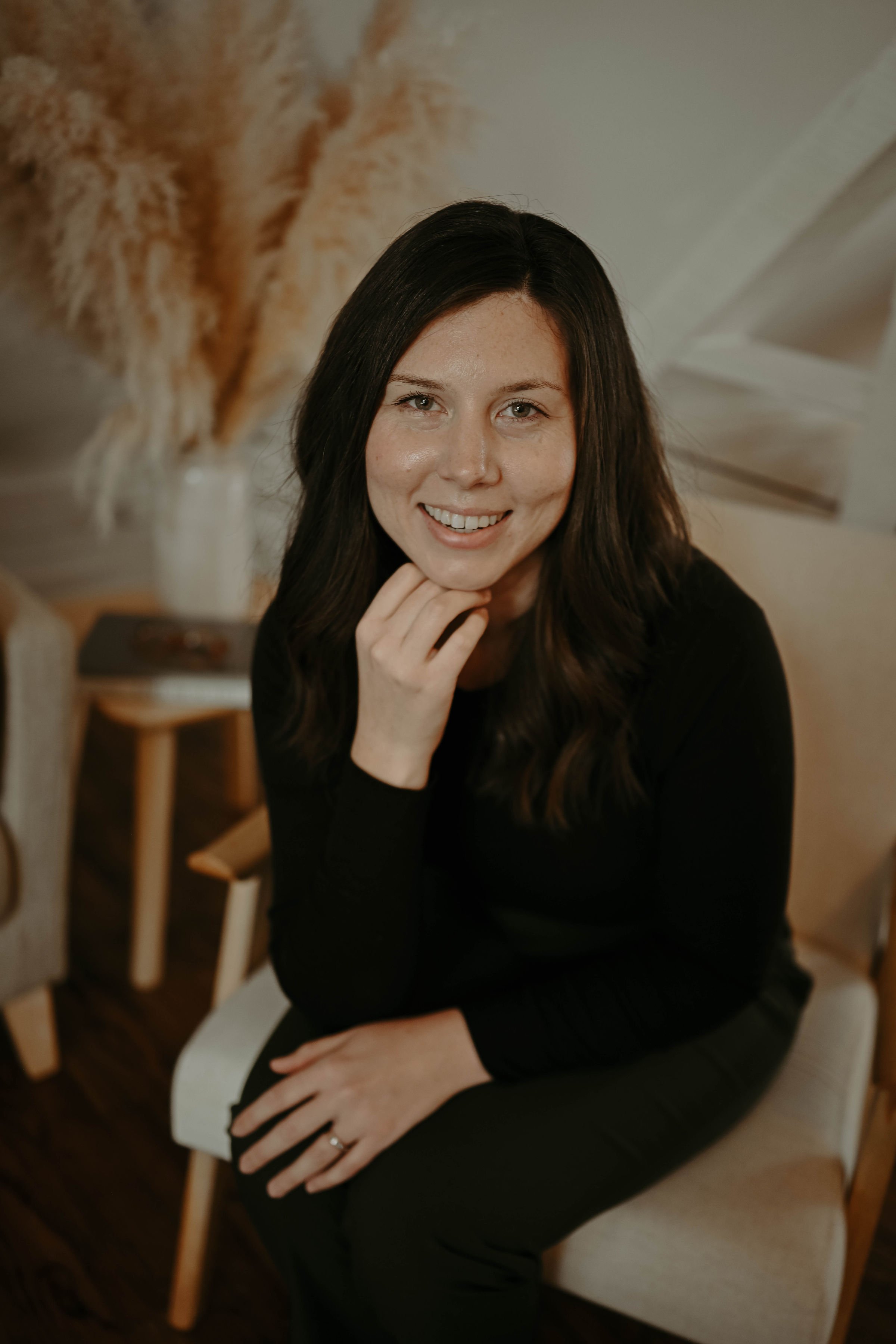 A woman with dark hair smiling and resting her chin on her hand, sitting on a light-colored chair indoors.