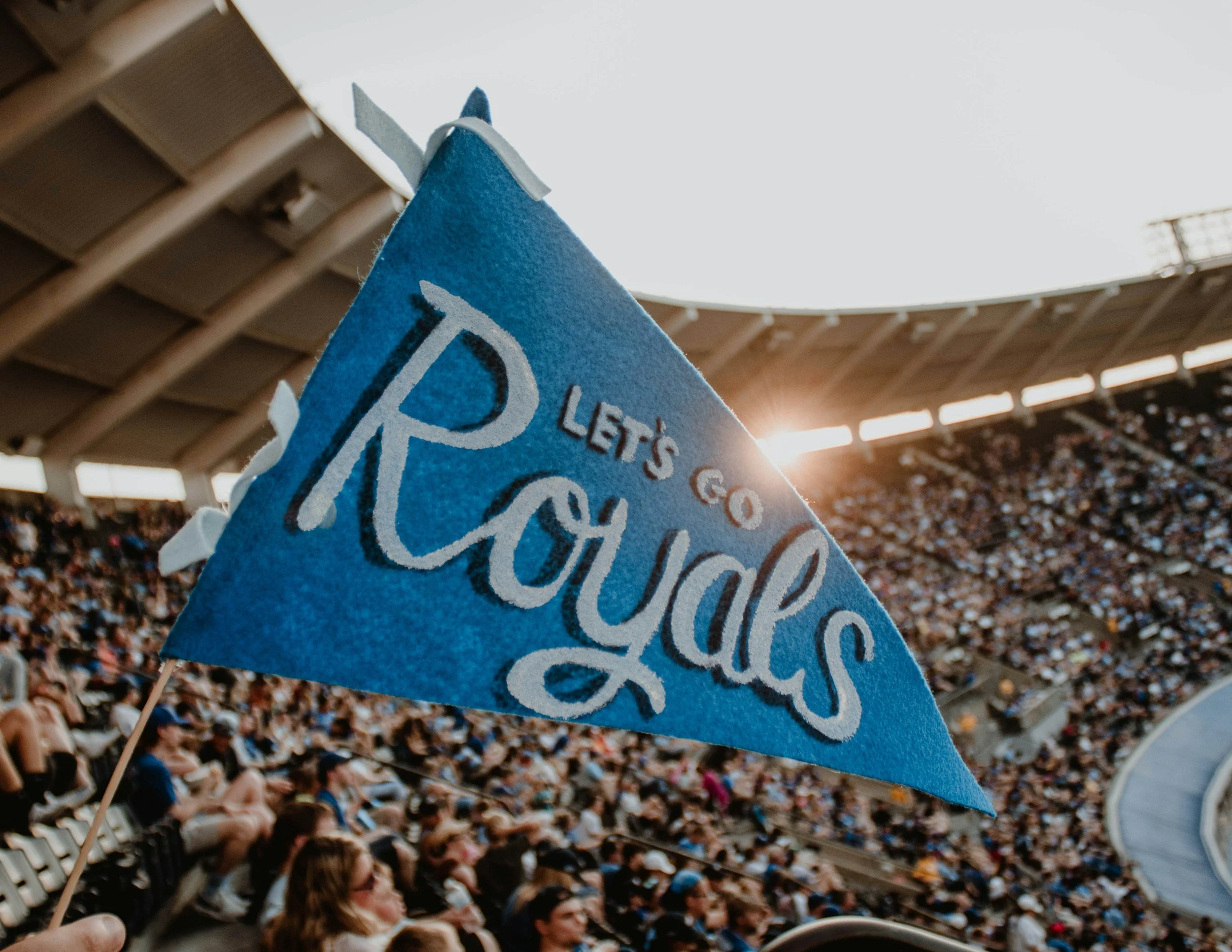 A blue protest or rally flag with partially visible white text, held above a large crowd of seated people in an outdoor stadium with a wooden roof structure and sunlight in the background.