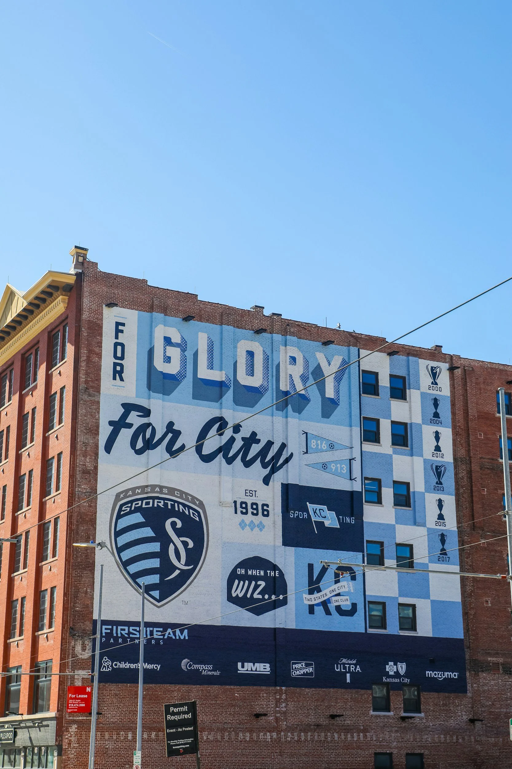 Large mural on side of a building celebrating Sporting Kansas City, displaying the team's name, logo, timeline of championships from 2000 to 2017, and various sponsor logos at the bottom.