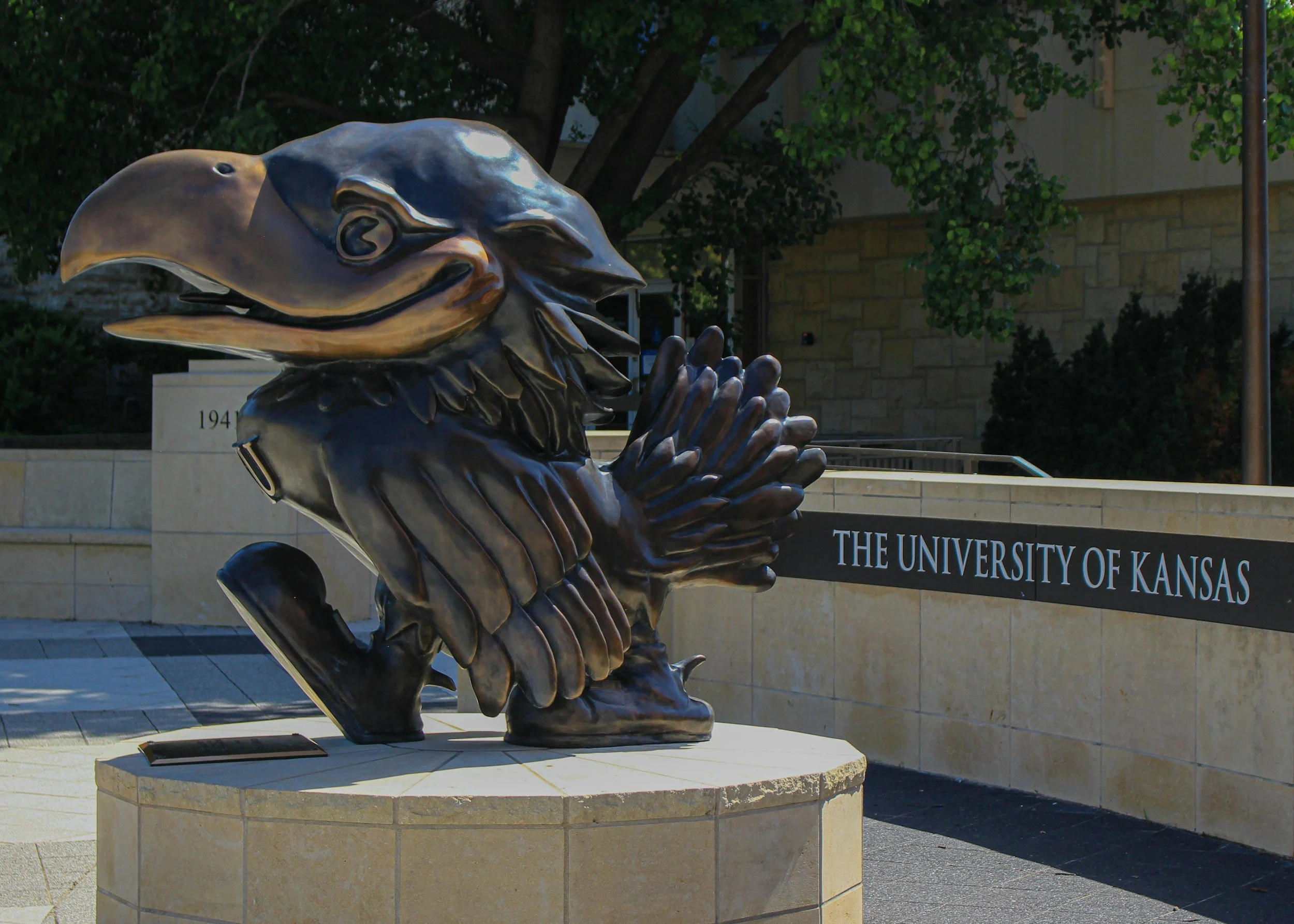 Bronze statue of a bird with its beak open, positioned outdoors near a building with trees in the background, and a sign for the University of Kansas in the background.