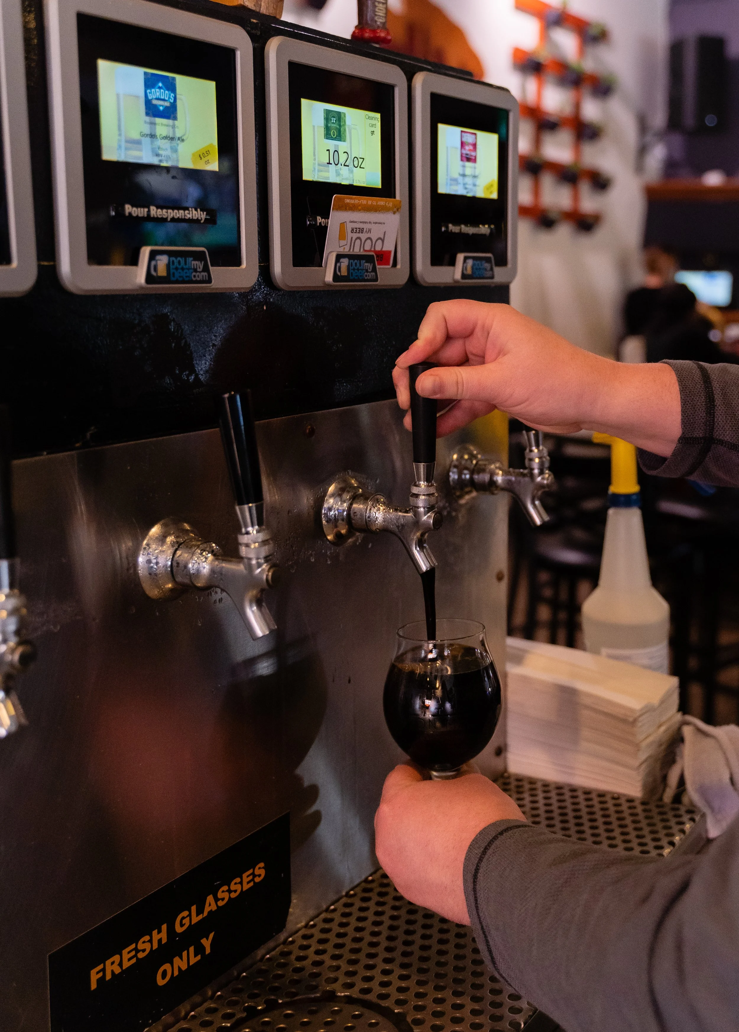 A person is filling a glass of dark beer from a tap at a bar or brewery. The taps are part of a self-service beer station with digital screens showing different beer options, including Gordo's Golden Ale. The person is wearing a gray long-sleeve shirt, and there are shelves with bottles and other bar equipment in the background.