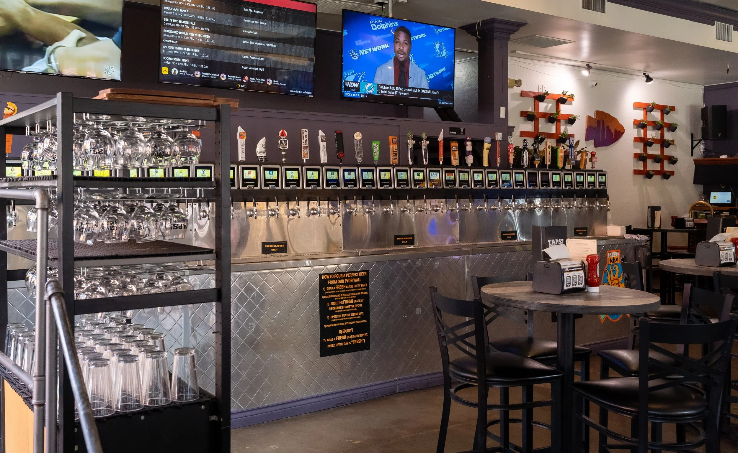 Inside a sports bar or restaurant with multiple tap beer dispensers, a TV screen showing a news broadcast with a man in a suit, and another TV screen displaying a menu or information. There are tables with chairs, glassware, and some decorative wall art.