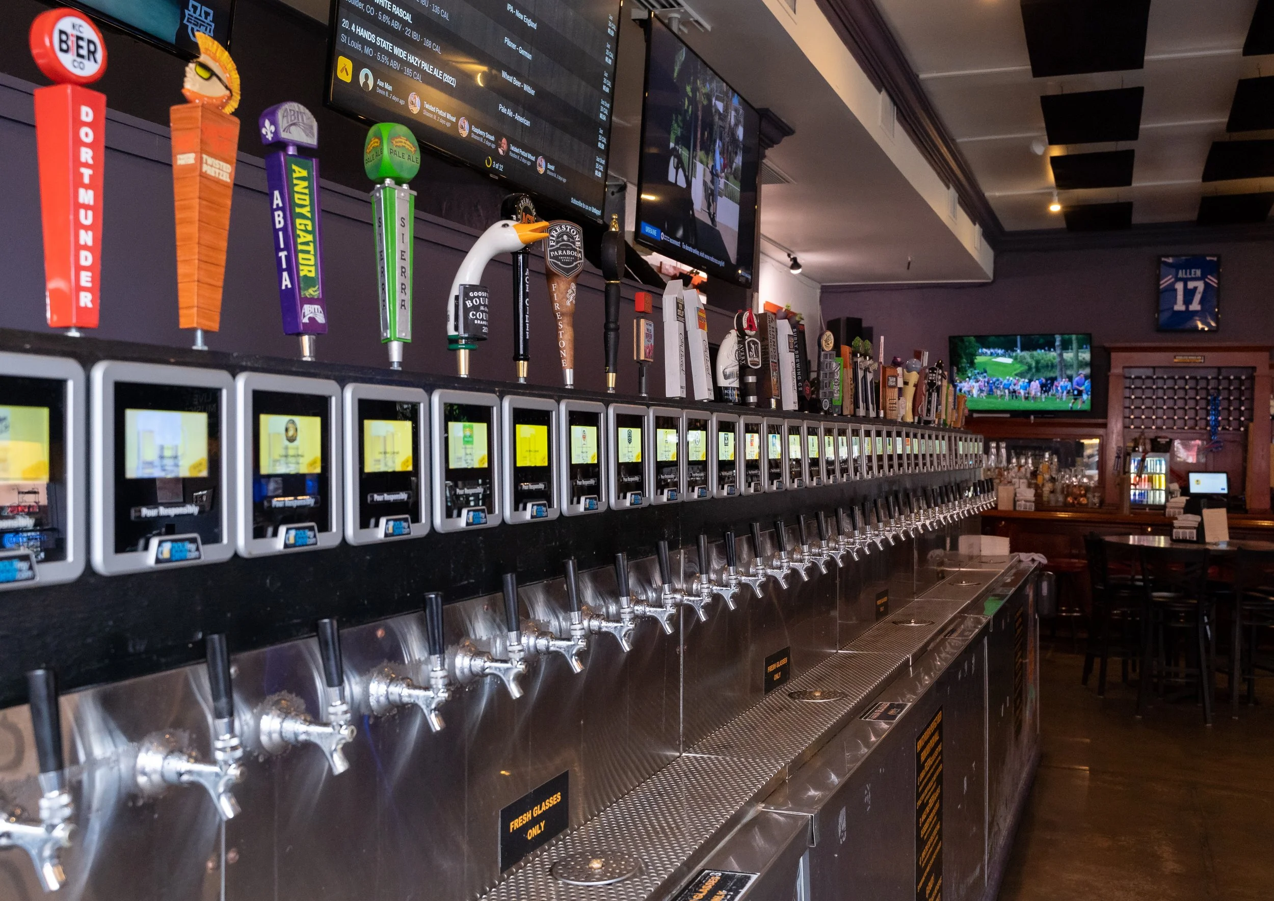 A bar with numerous beer taps with colorful handles, digital menu screens overhead, and a television displaying a golf game in the background.
